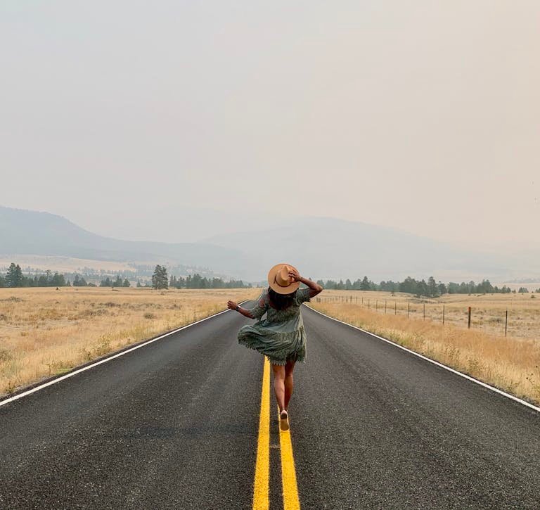 A woman in a dress with a hat walks on a deserted country road in Helena, MT, USA, exuding wanderlust.