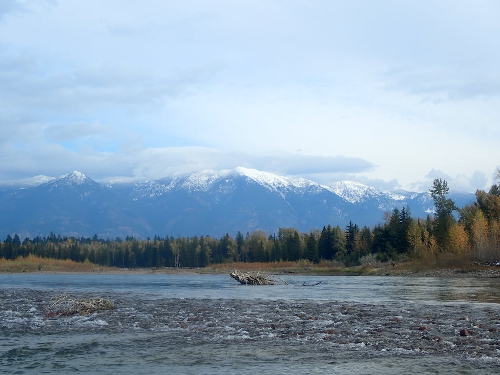 Peaceful view of Whitefish River with snow-capped mountains and autumn forest in Whitefish, Montana.