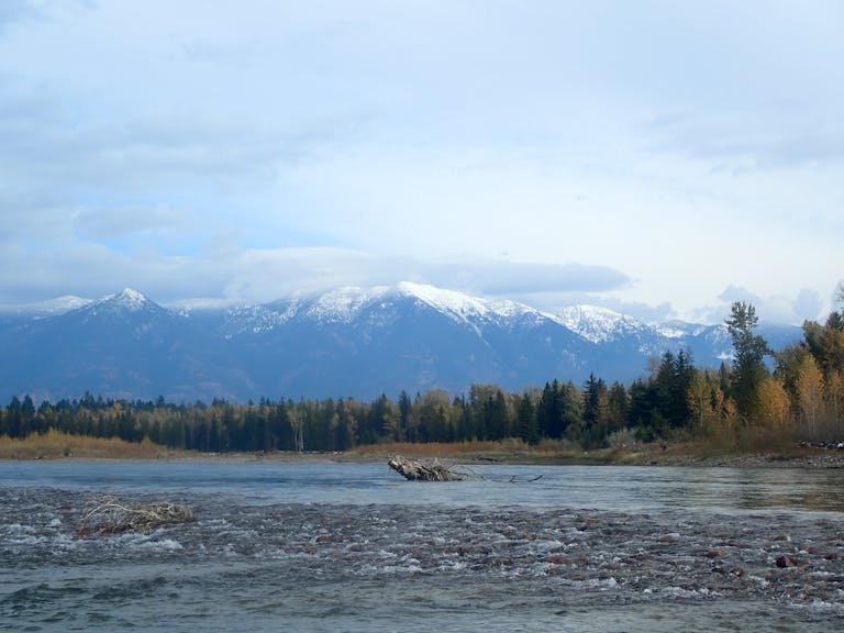 Peaceful view of Whitefish River with snow-capped mountains and autumn forest in Whitefish, Montana.