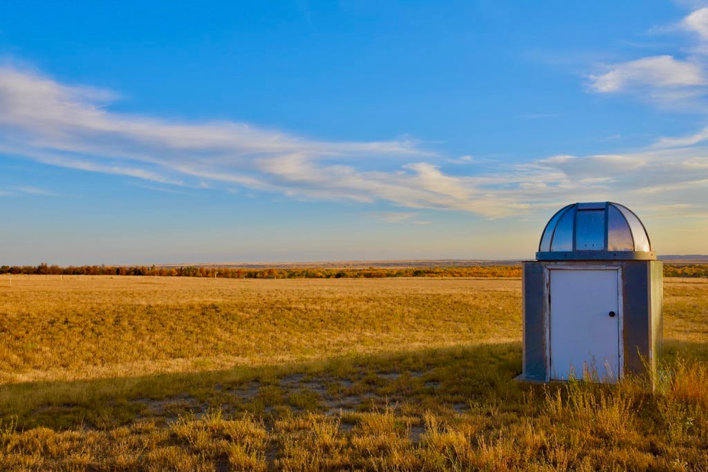A small weather observatory building placed in a vast open grassland under a clear blue sky in Glasgow, Montana.