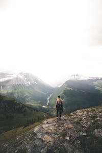 Back view of unrecognizable young female hiker with long blond hair in casual clothes and backpack recreating on rocky hill slope and admiring spectacular mountain view on cloudy day
