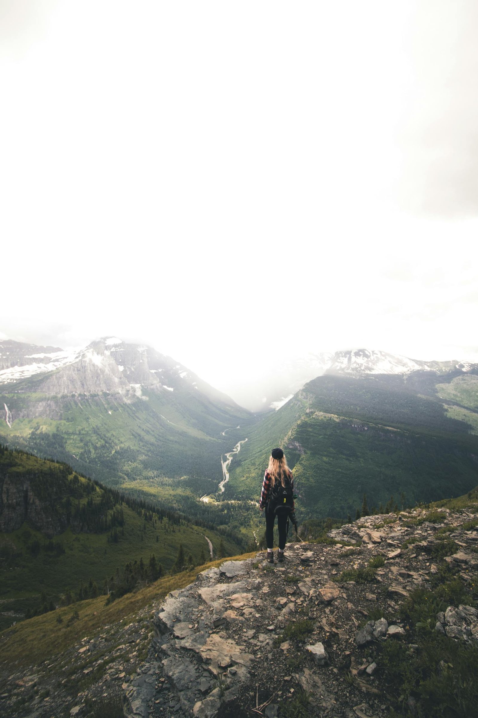 Back view of unrecognizable young female hiker with long blond hair in casual clothes and backpack recreating on rocky hill slope and admiring spectacular mountain view on cloudy day