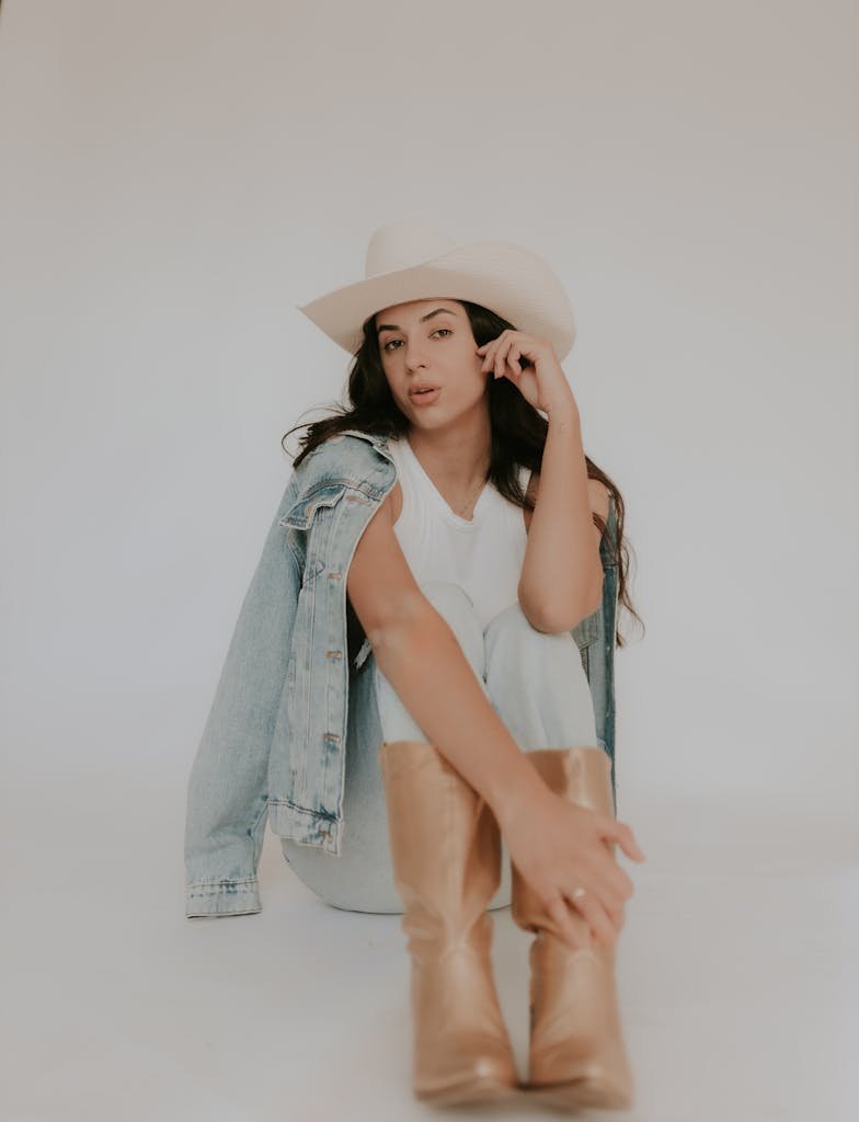 Young woman in cowboy hat and denim jacket posing stylishly in studio setting.